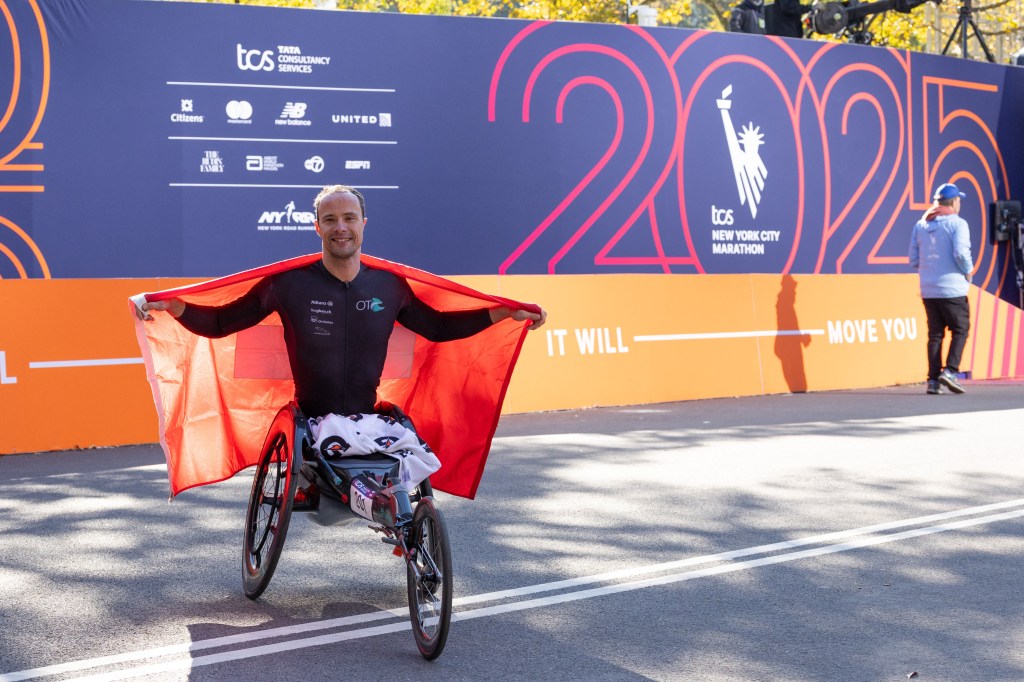 Marcel Hug in a racing wheelchair, holding a red flag and smiling, after winning the 2025 New York City Marathon.