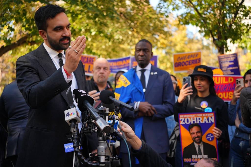 Zohran Mamdani speaks at a canvass launch in Harlem, with microphones from various news outlets in the foreground.