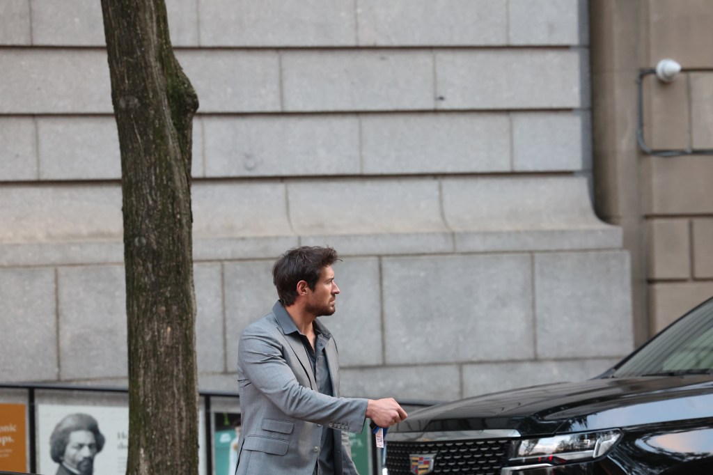 Bari Weiss’ bodyguard walking on the street next to a black Cadillac.