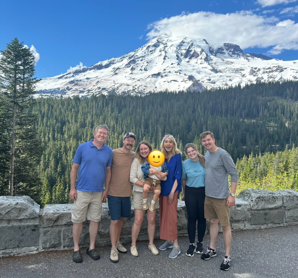 Six people and one baby with a blurred-out face smile while standing in front of a snow-capped mountain and evergreen trees.