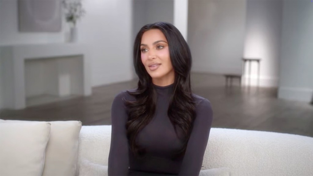 Kim Kardashian in a black long-sleeved shirt sitting on a white sofa.