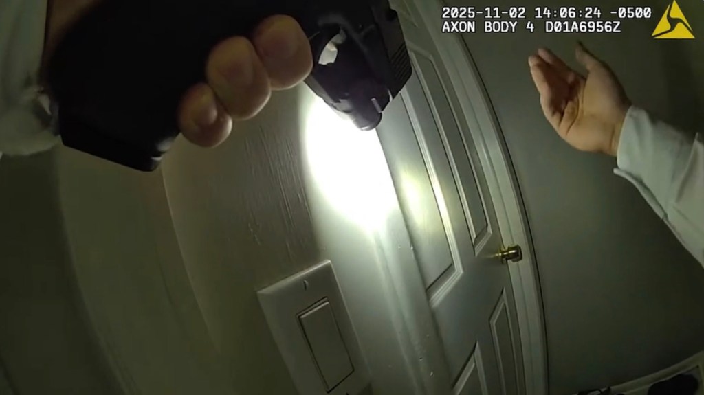 Body camera footage showing a gun pointed at a closed door, with a hand visible at the right.