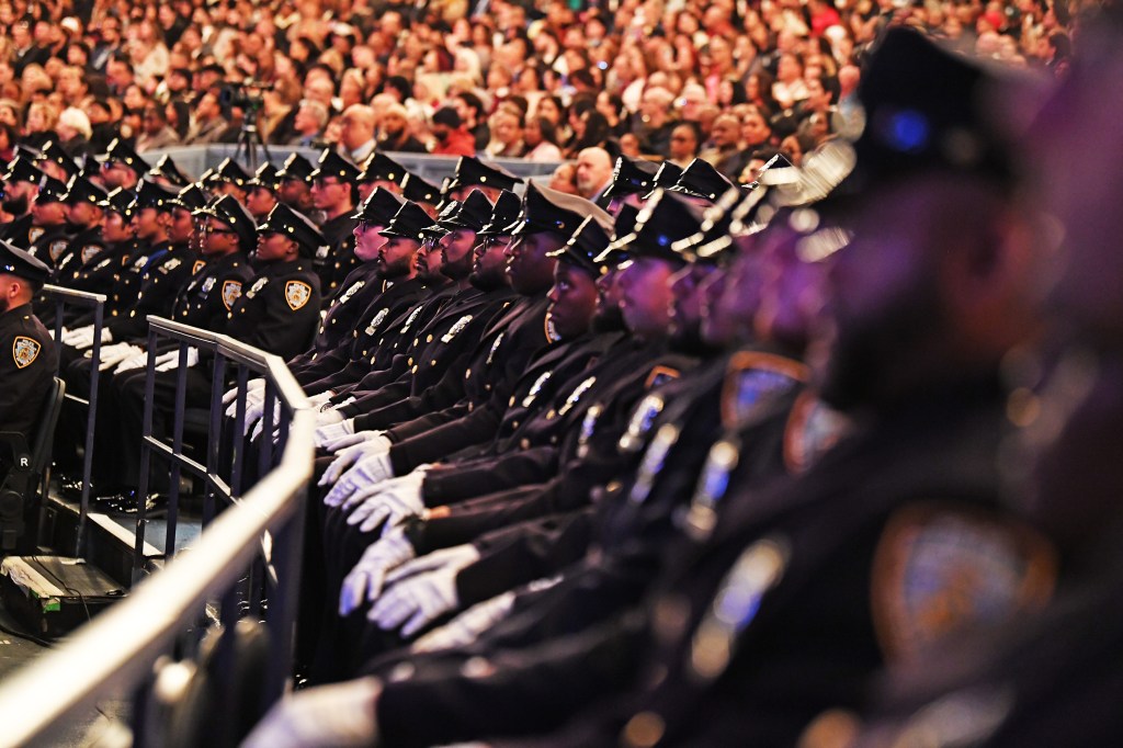 NYPD recruits in uniform and white gloves seated during a graduation ceremony, with a crowd visible in the background.