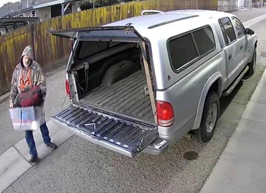 A man carrying a red bag and a clear bin of items walks away from the open tailgate of a silver pickup truck.