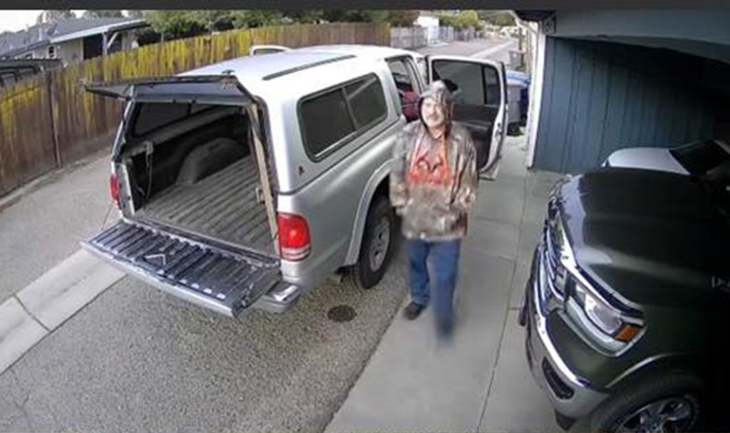 Surveillance footage showing Ron Dailey wearing a camouflage jacket and jeans, standing near a silver pickup truck with its tailgate down.