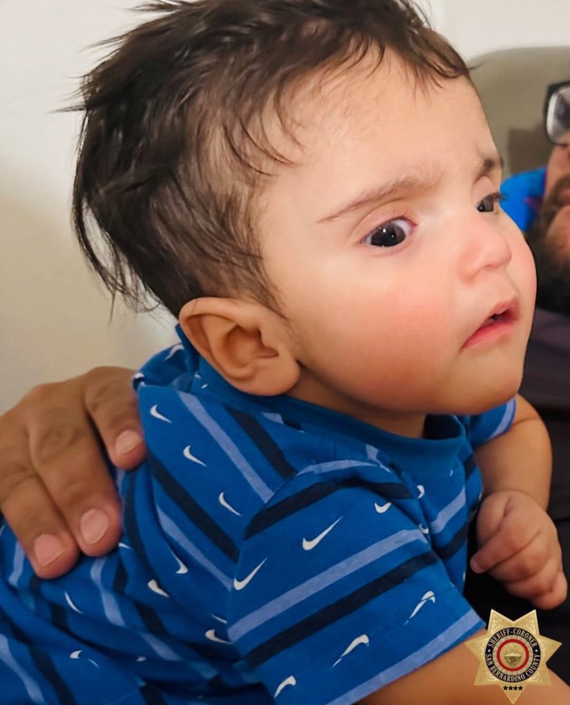 A 7-month-old baby, Emmanuel Haro, with dark hair and wide eyes, looks right while being held, wearing a blue striped shirt with white Nike swooshes.