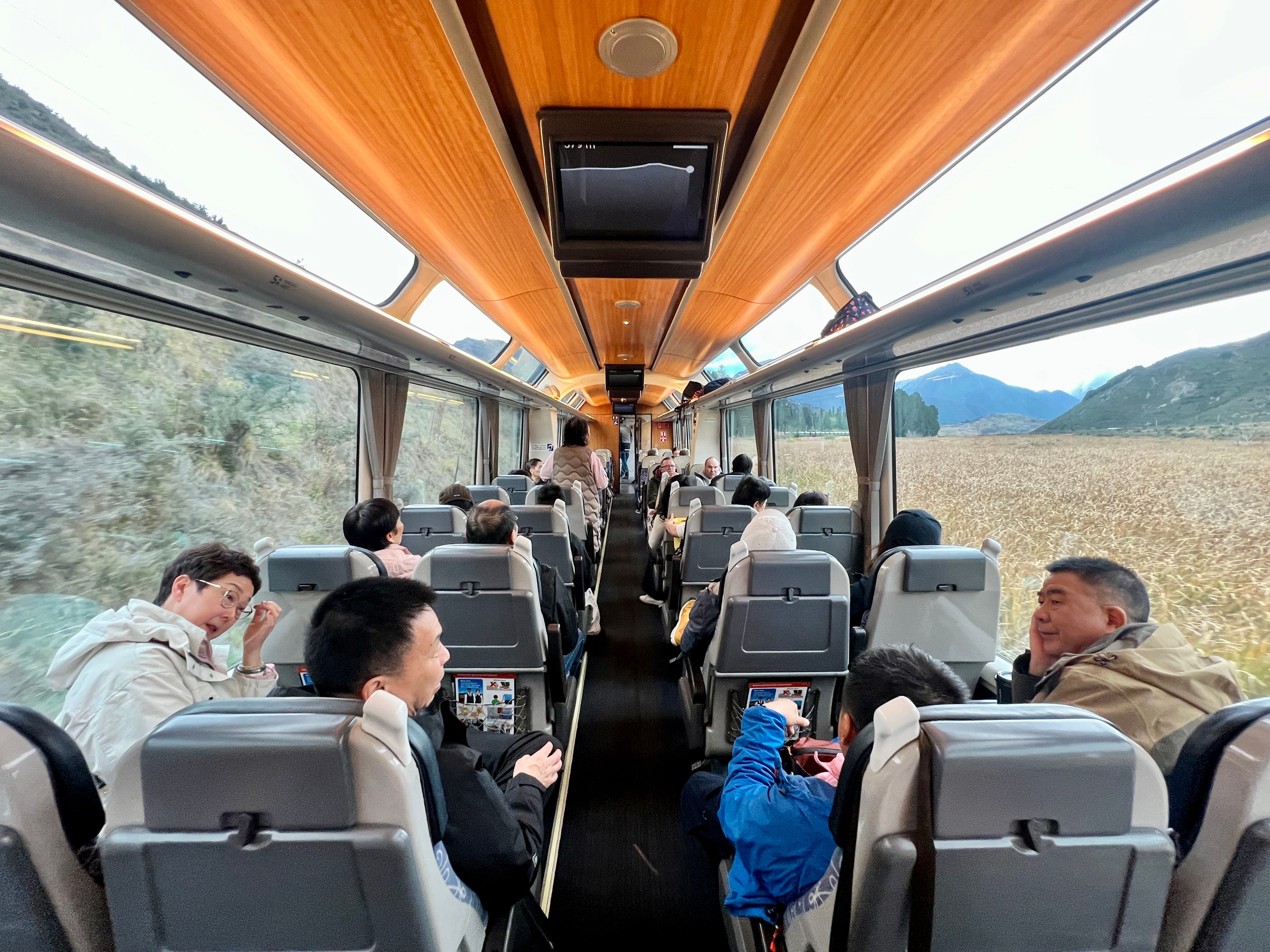 View of train car from back, interior - two seats on each side of aisle, wood-like ceilings, screens hanging from them