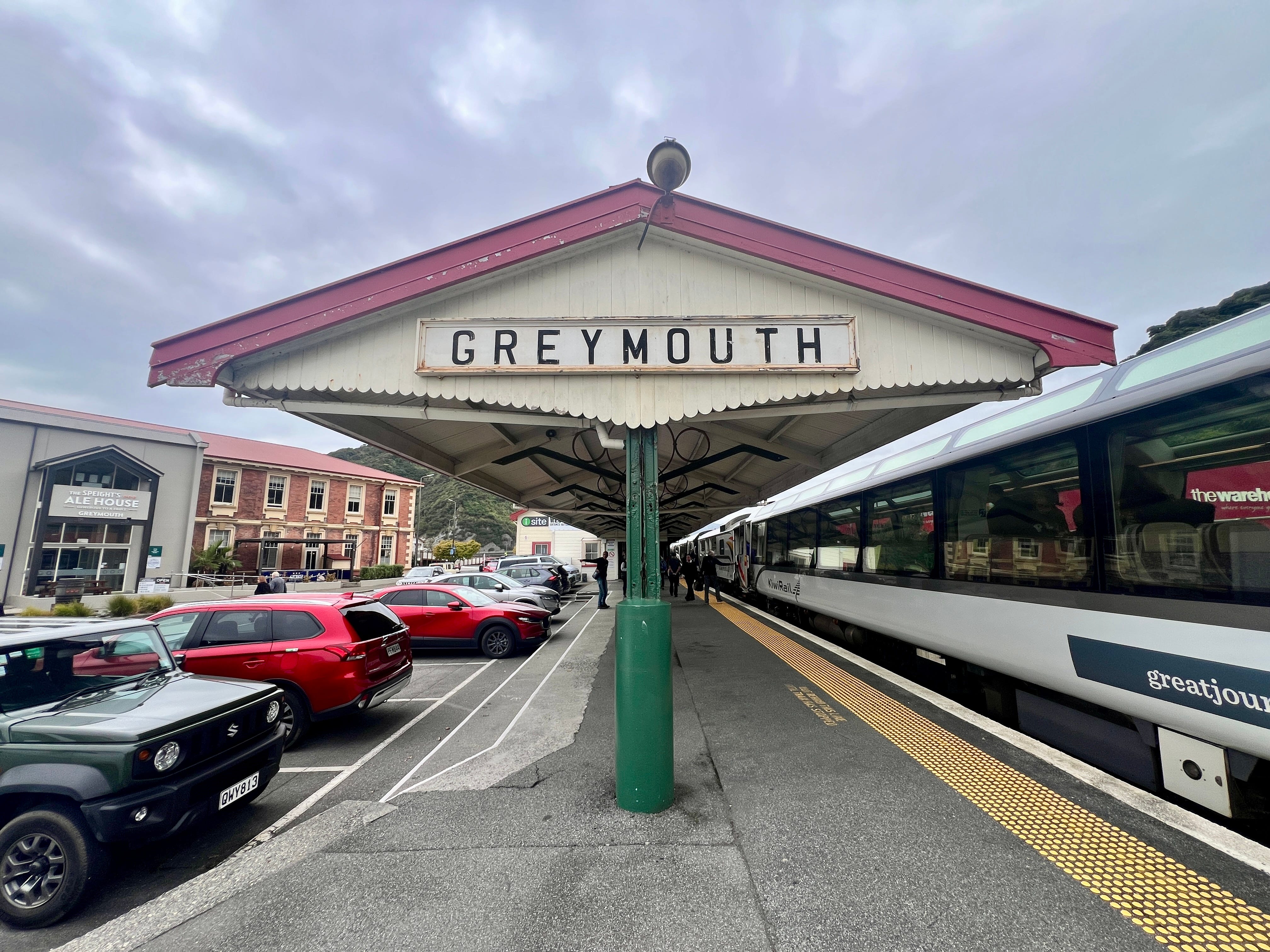 Greymouth train station sign with train, parked cars on either side of it