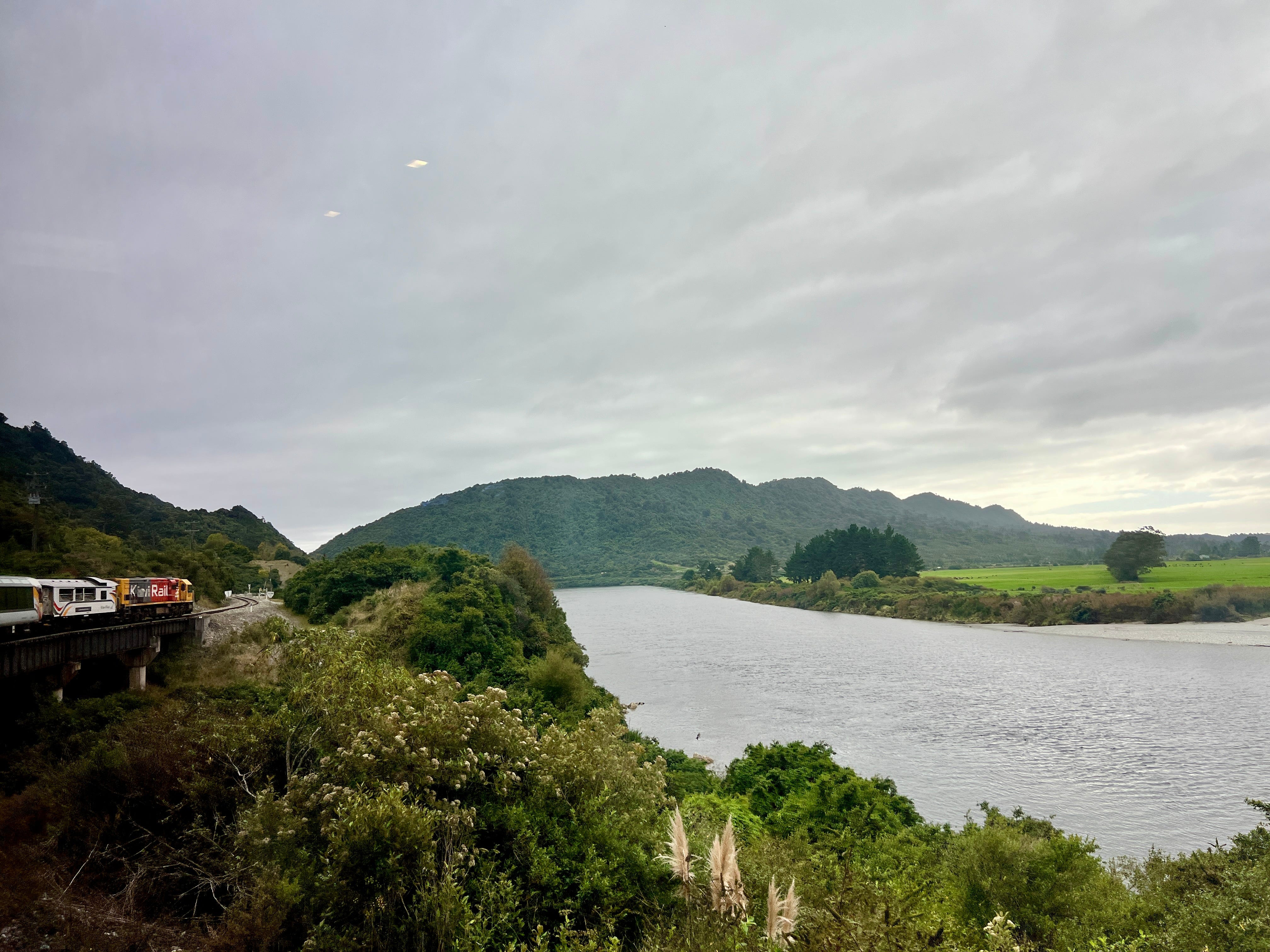 View of train on tracks next to body of water, mountains in New Zealand