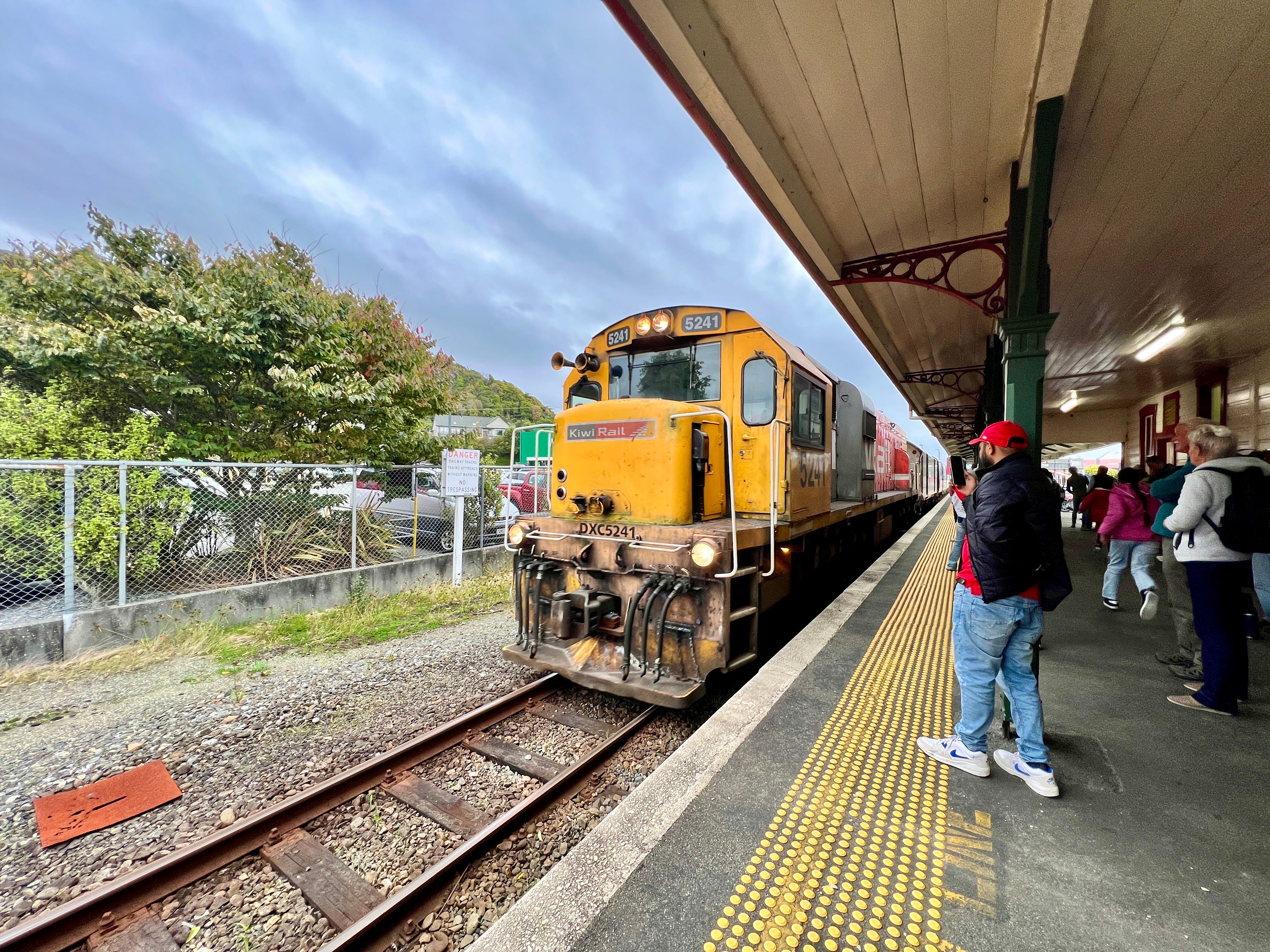 Yellow Kiwi Rail train pulling up to station as people wait nearby