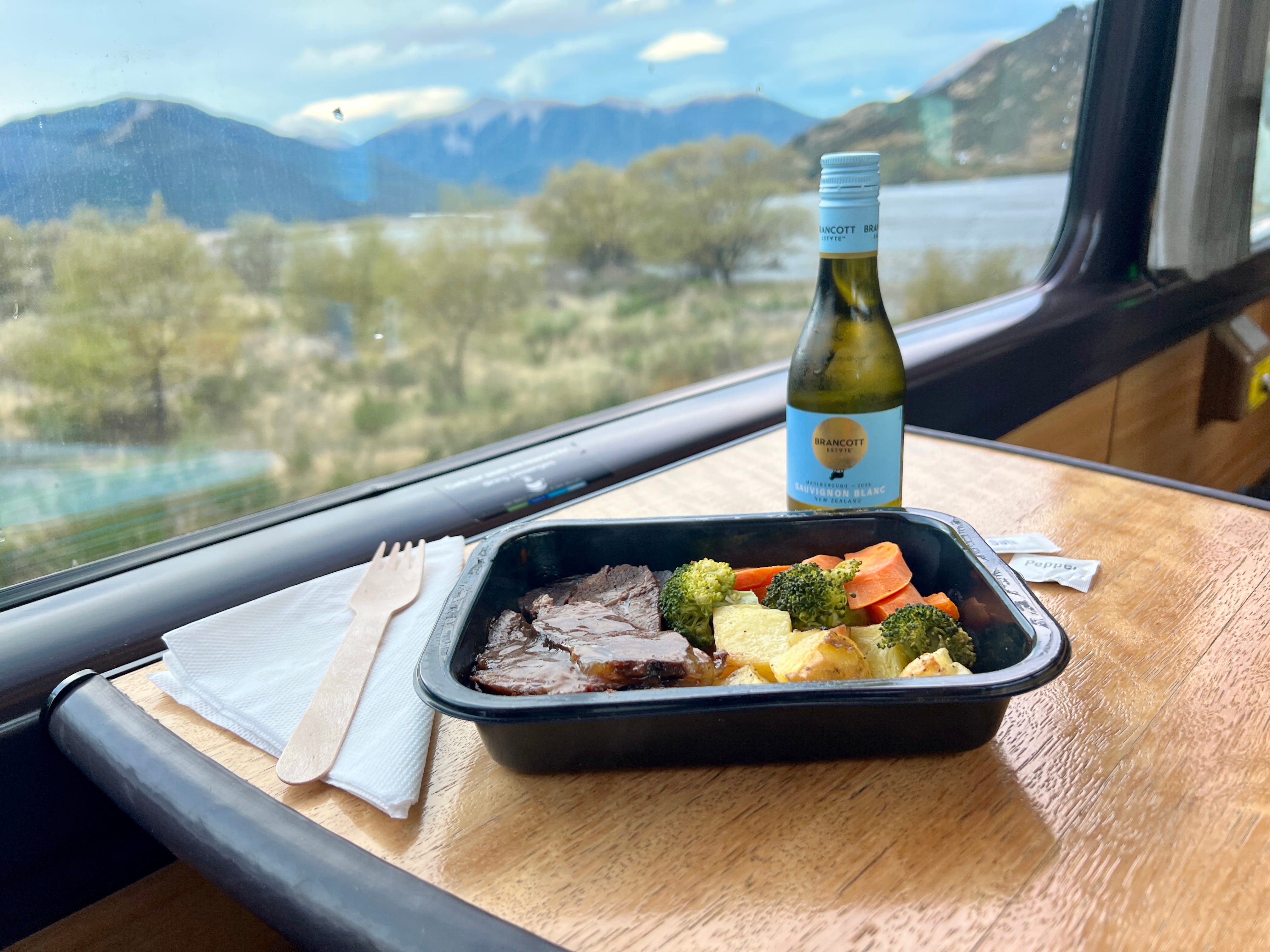 View of plastic container with veggies, meat, next to small bottle of wine on small tray table