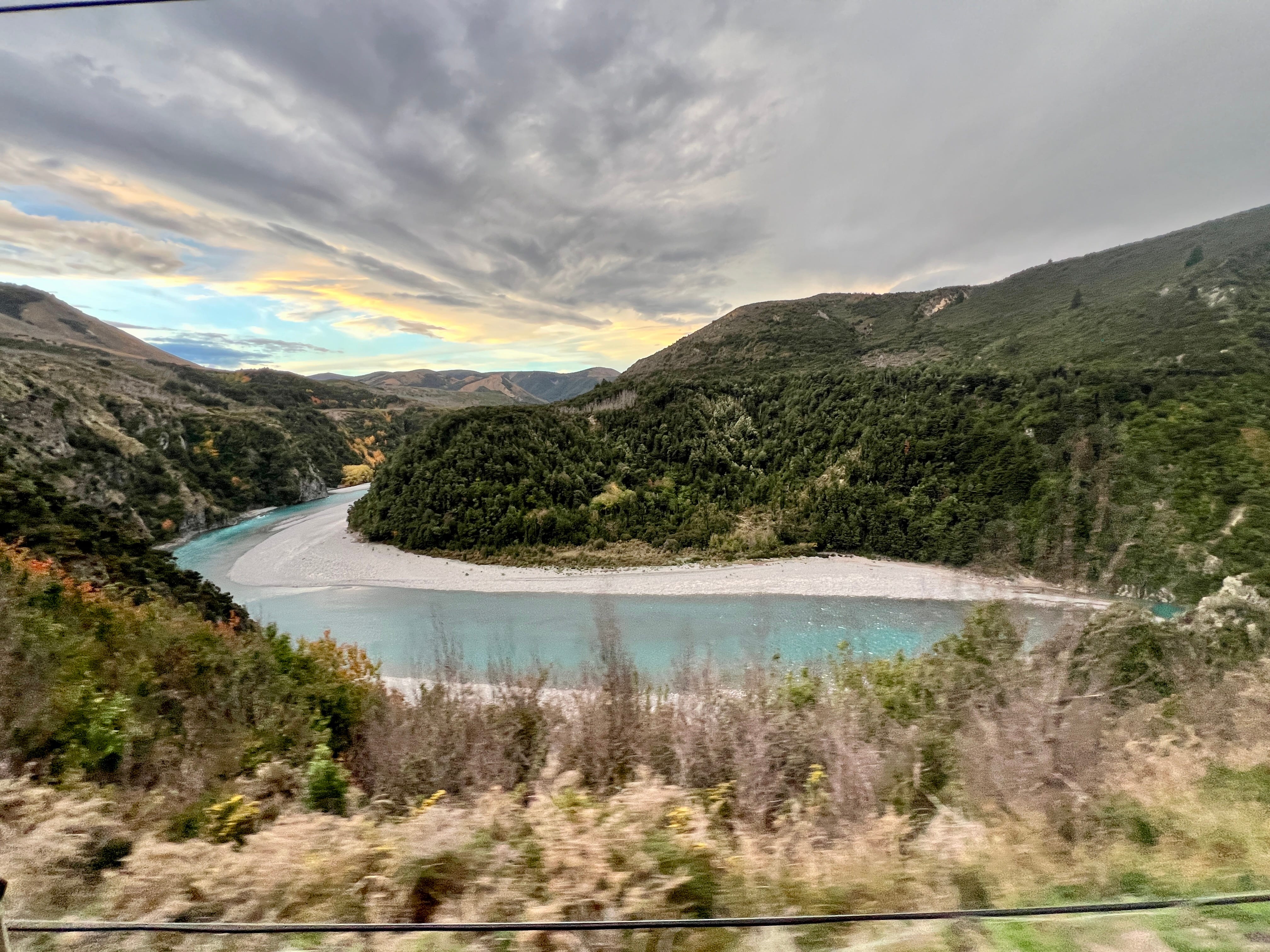View of body of water, clouds, tree-covered mountains from TranzAlpine train window