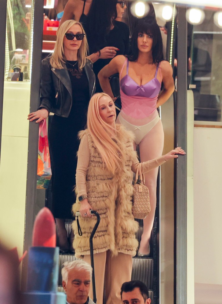 Bianca Censori in a lilac and pink bodysuit, with Alexandra Censori in a black midi dress and leather jacket, and another relative in a faux fur coat, on an escalator.