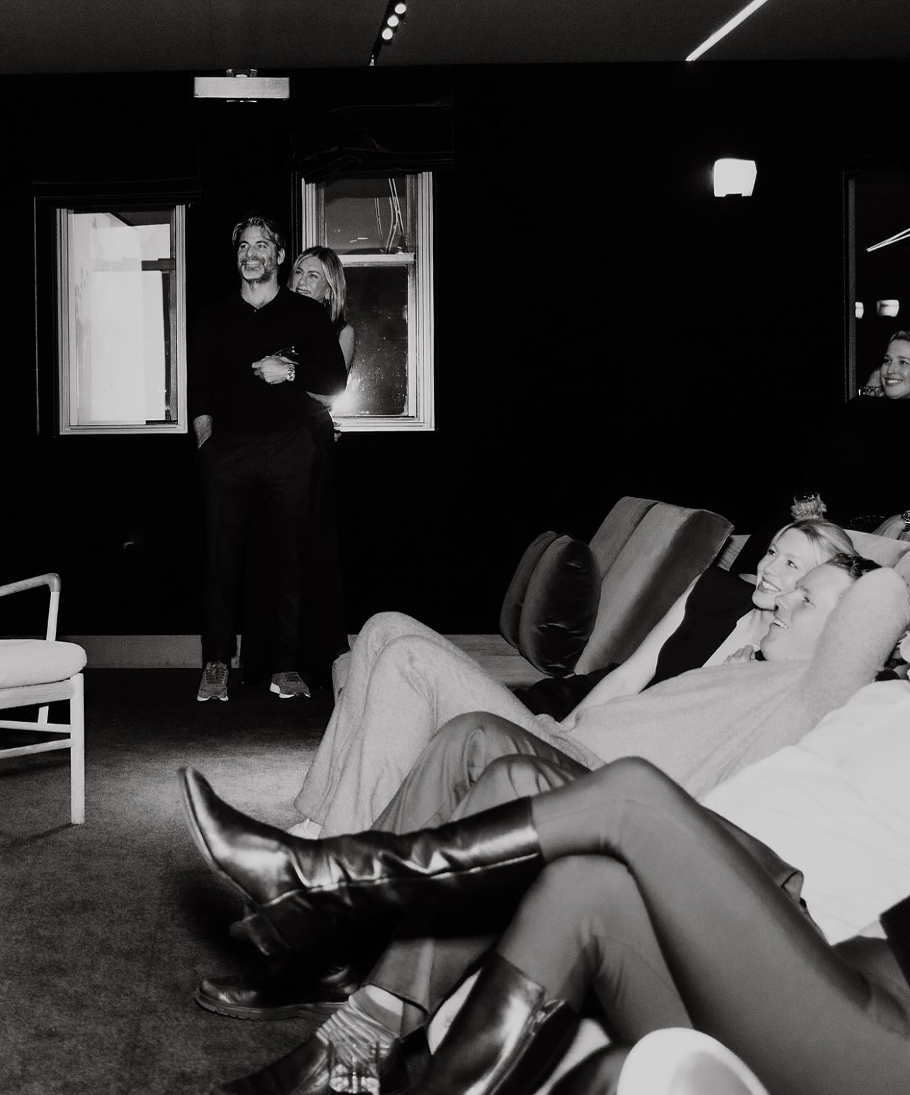 Jennifer Aniston hugging Jim Curtis from behind, while other guests relax on a couch.