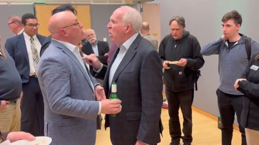 Thomas Speciale II confronts John Brennan, pointing his finger at the former CIA Director while holding a green bottle.