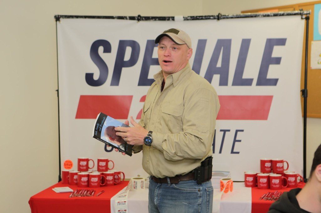 Thomas Speciale, a 20-year veteran intel analyst, holds a book in front of a campaign banner.