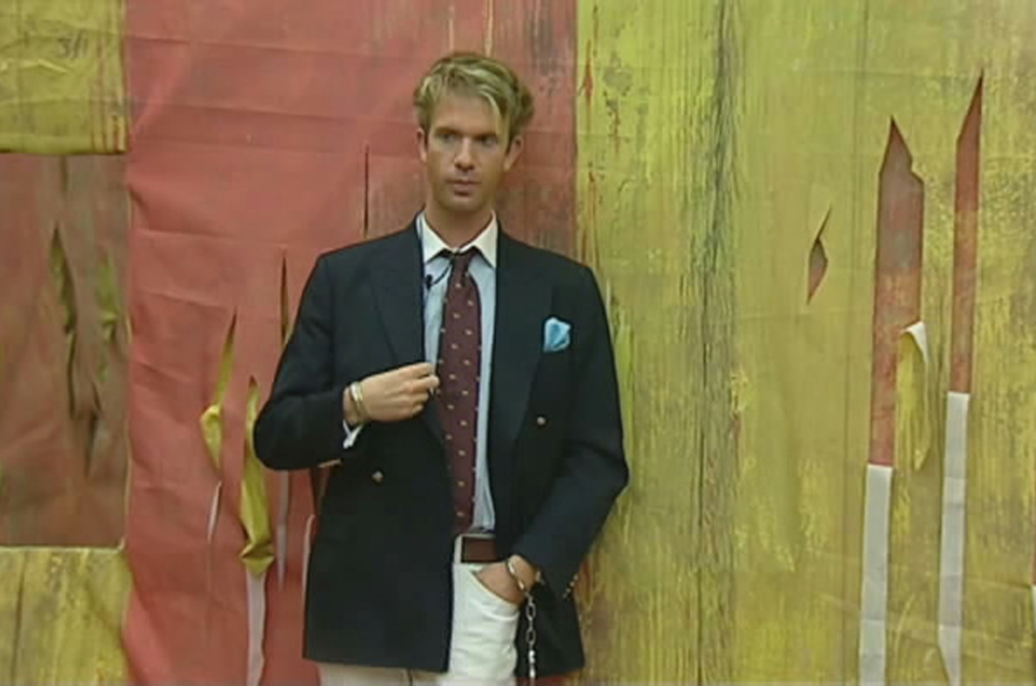 A blonde man in a suit and tie standing against a colorful, peeling wall.