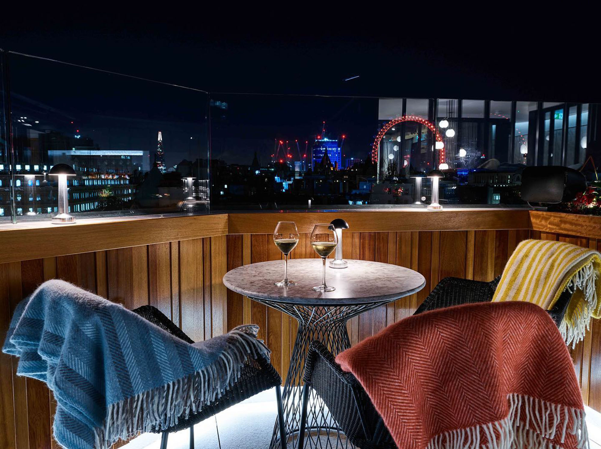 Rooftop bar at night with a small table, two chairs with blankets, and city lights in the background including the London Eye.