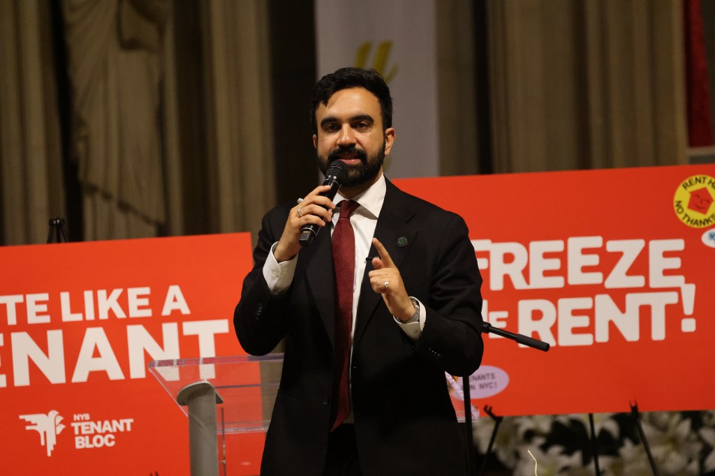 Zohran Mamdani speaking at a Tenants' Rights Rally with a red banner behind him that reads