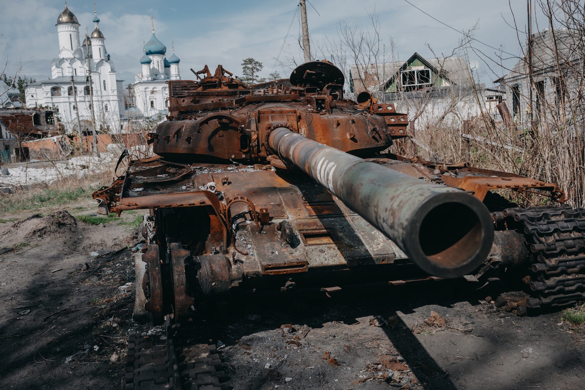 A rust-coloured damaged tank on muddy ground in front of some small buildings and what appaers to be an Orthodox church