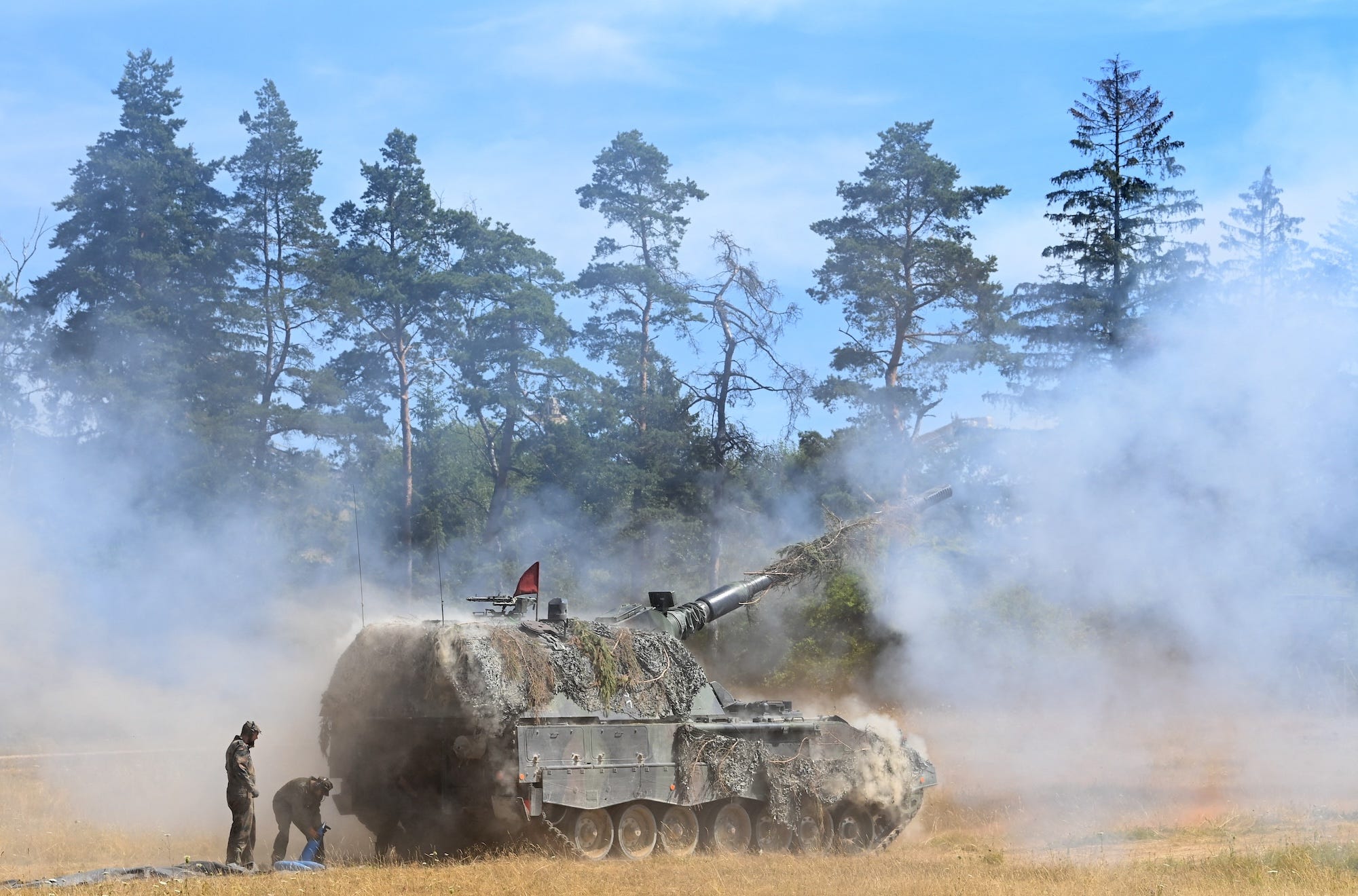A large, wheeled piece of weaponry with a large turret and covered in camouflage netting with smoke around it, with two figures standing beside it and trees and a blue sky in the background