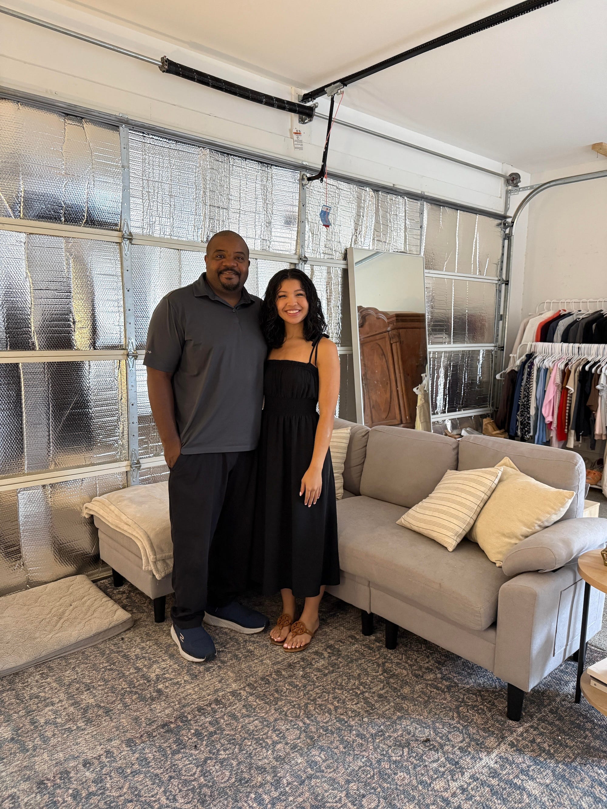 A dad and his daughter stand together in front of a couch in a garage.