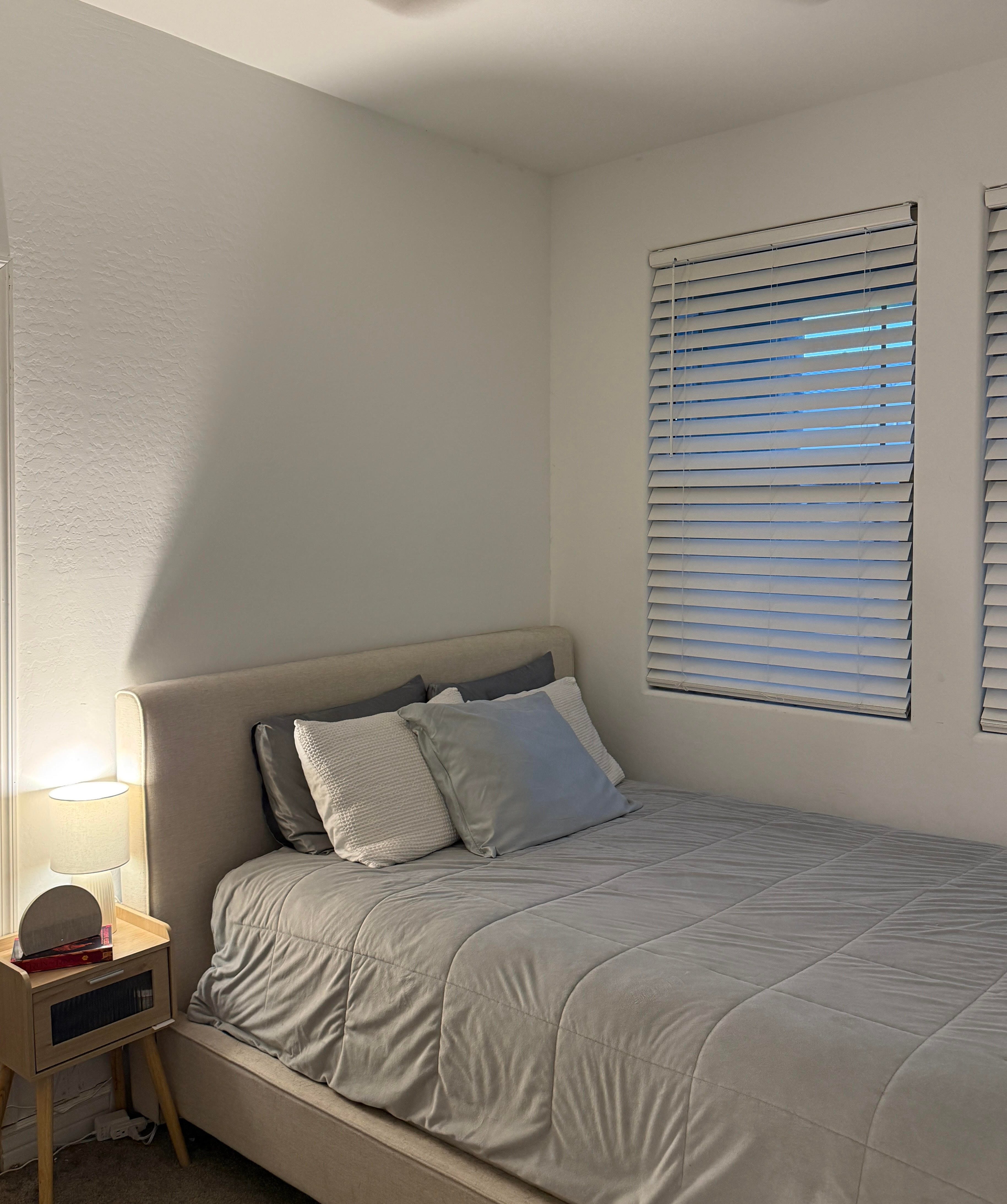 A bedroom with a bed against the wall. A small nightstand sits next to it, and there are two windows on the wall.