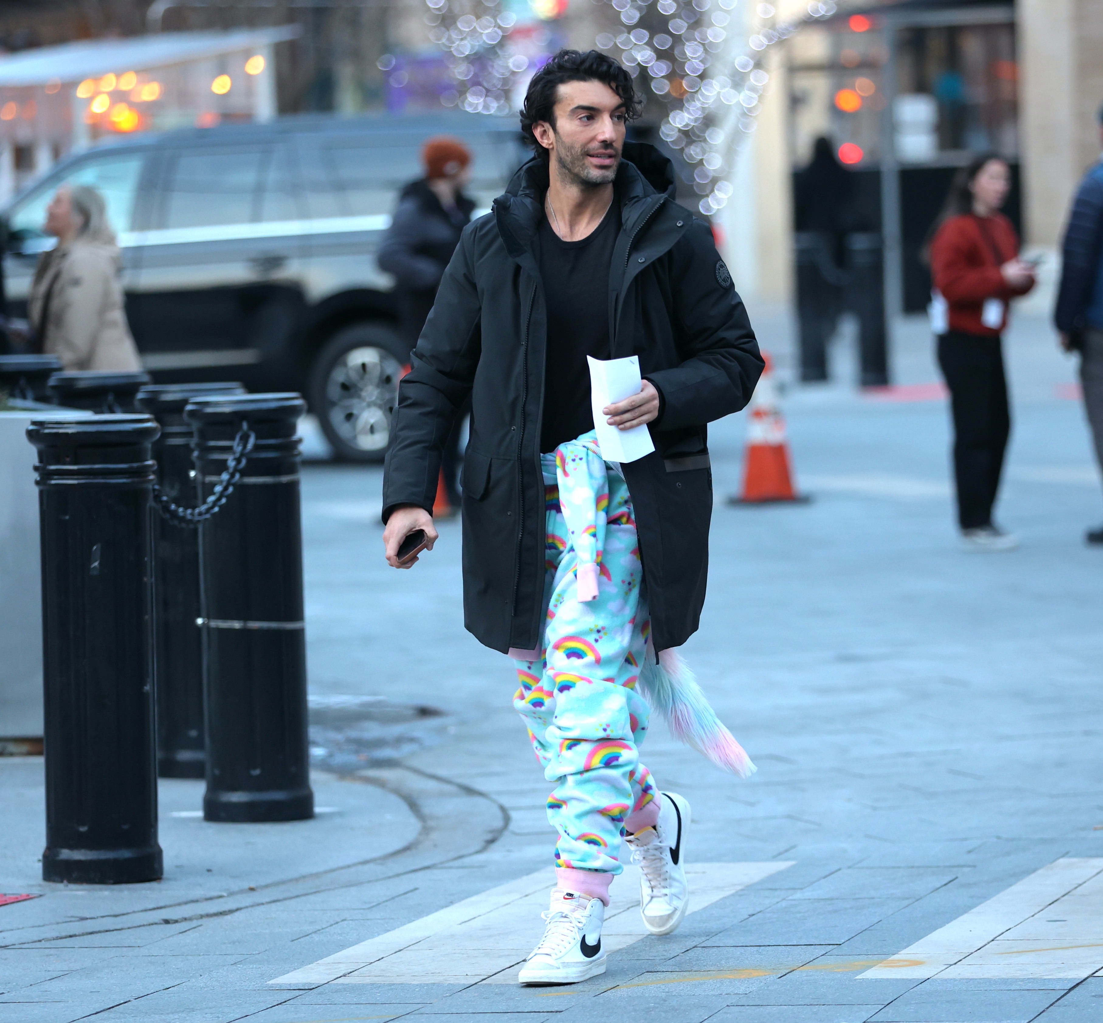 Justin Baldoni wearing a black coat, rainbow-patterned pants, and white sneakers while walking on set in New Jersey.