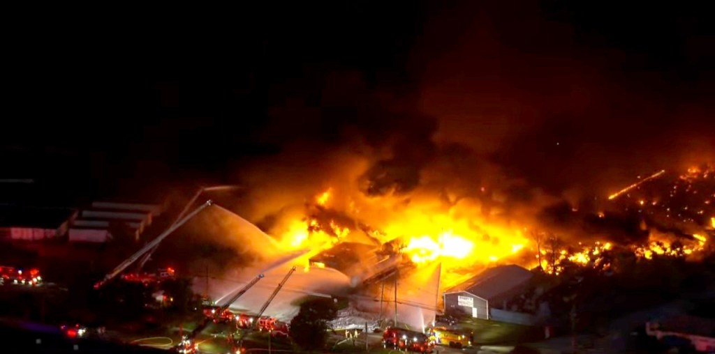 Aerial view of multiple buildings engulfed in a large fire at night, with several fire truck ladders spraying water.