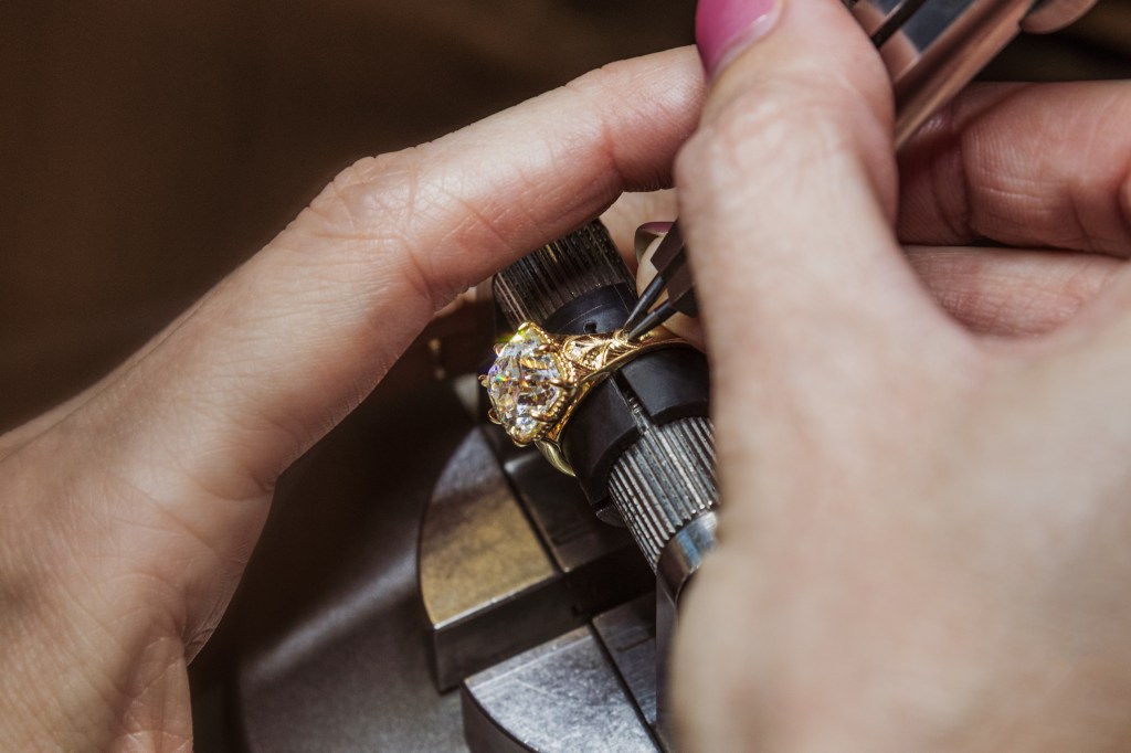 A hand holding tweezers working on a gold ring with a large diamond.