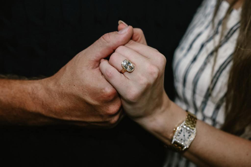 A woman wearing a large engagement ring and a gold watch holds hands with a man.