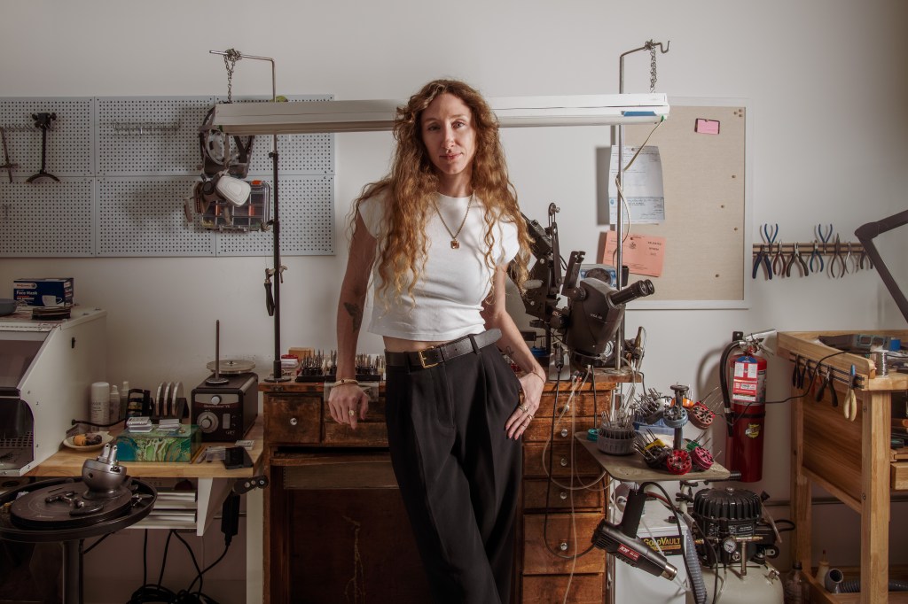 A woman with long wavy red hair and a white shirt with a golden necklace, leaning against a wooden workbench in a workshop.