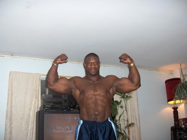 Bodybuilder Joseph Biggs flexing his biceps in a living room.