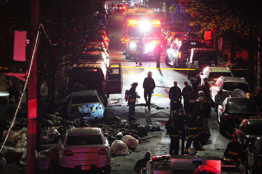Firefighters and emergency personnel responding to a car fire at night, with debris and a burnt-out car visible.