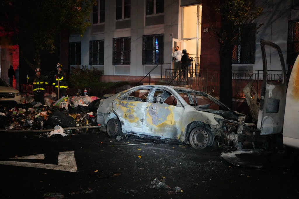 A burnt-out car with two firefighters standing near a pile of debris.