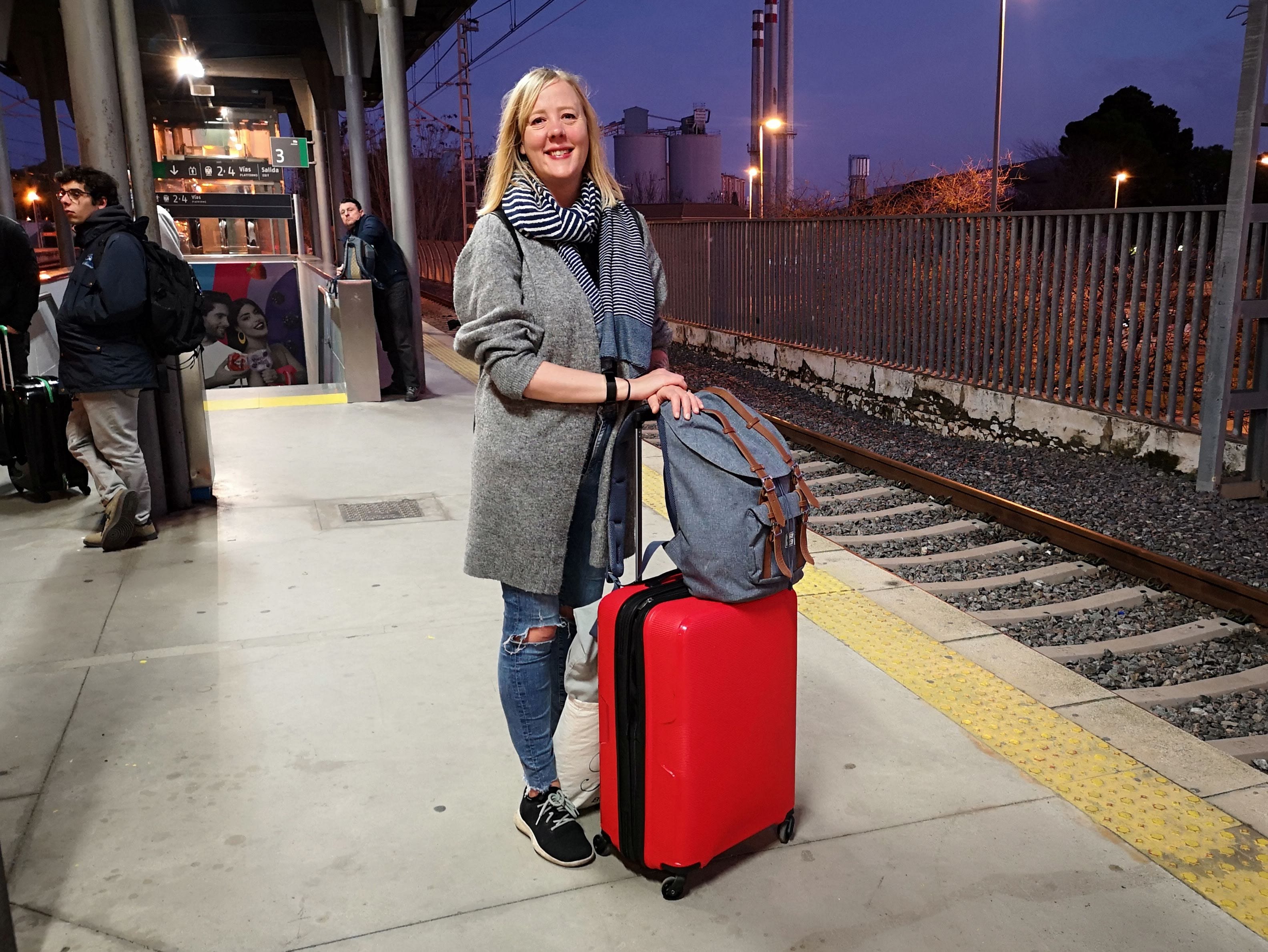 Charlie Brown standing on a train station platform holding the handle of a red suitcase with wheels.