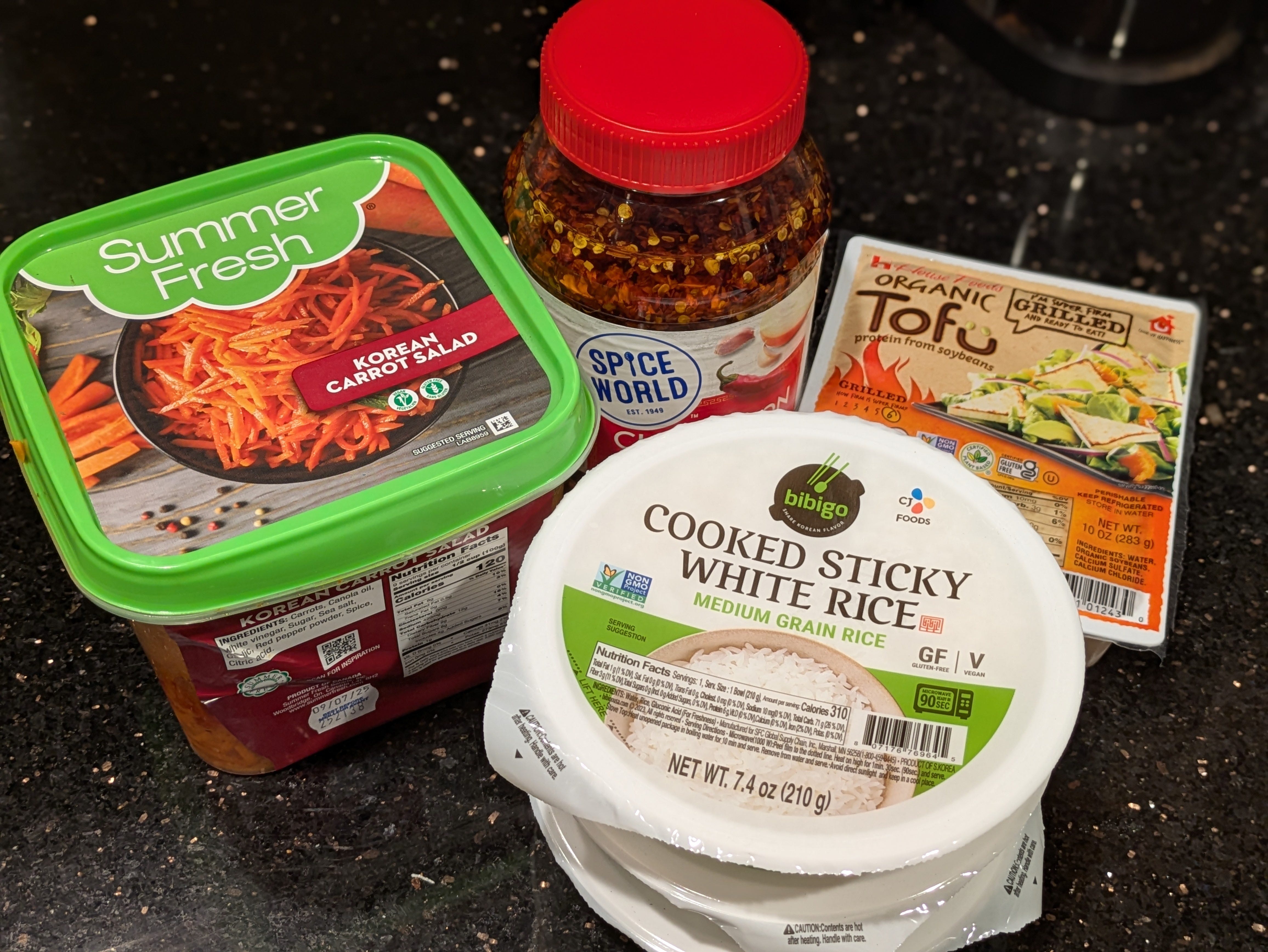 A collection of ingredients the author uses for rice bowls, including sticky white rice, organic tofu, Korean carrot salad, and Spice World chili onion crunch.