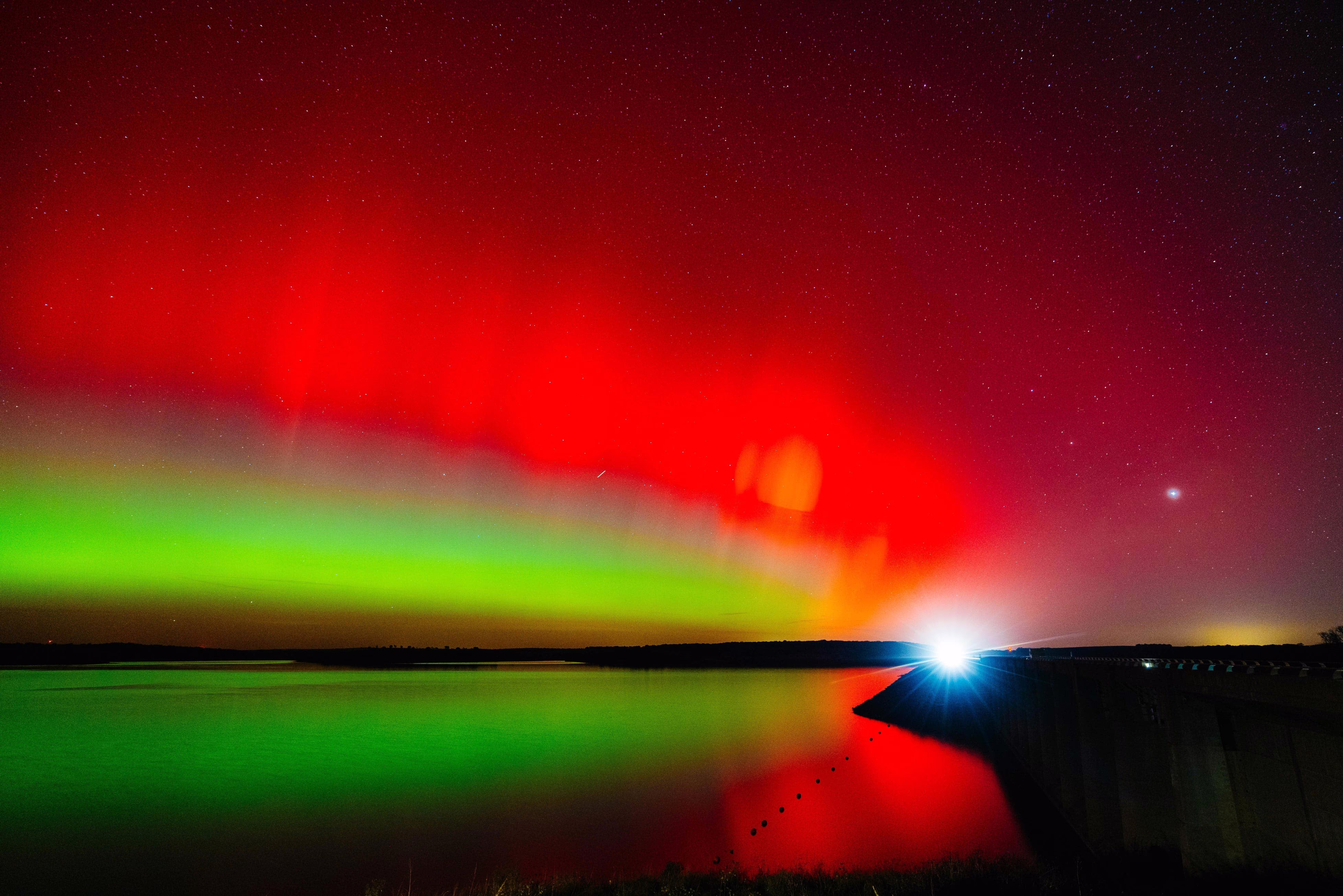 A night view of a distant car's headlights, and a red-and-green-colored aurora in the sky.