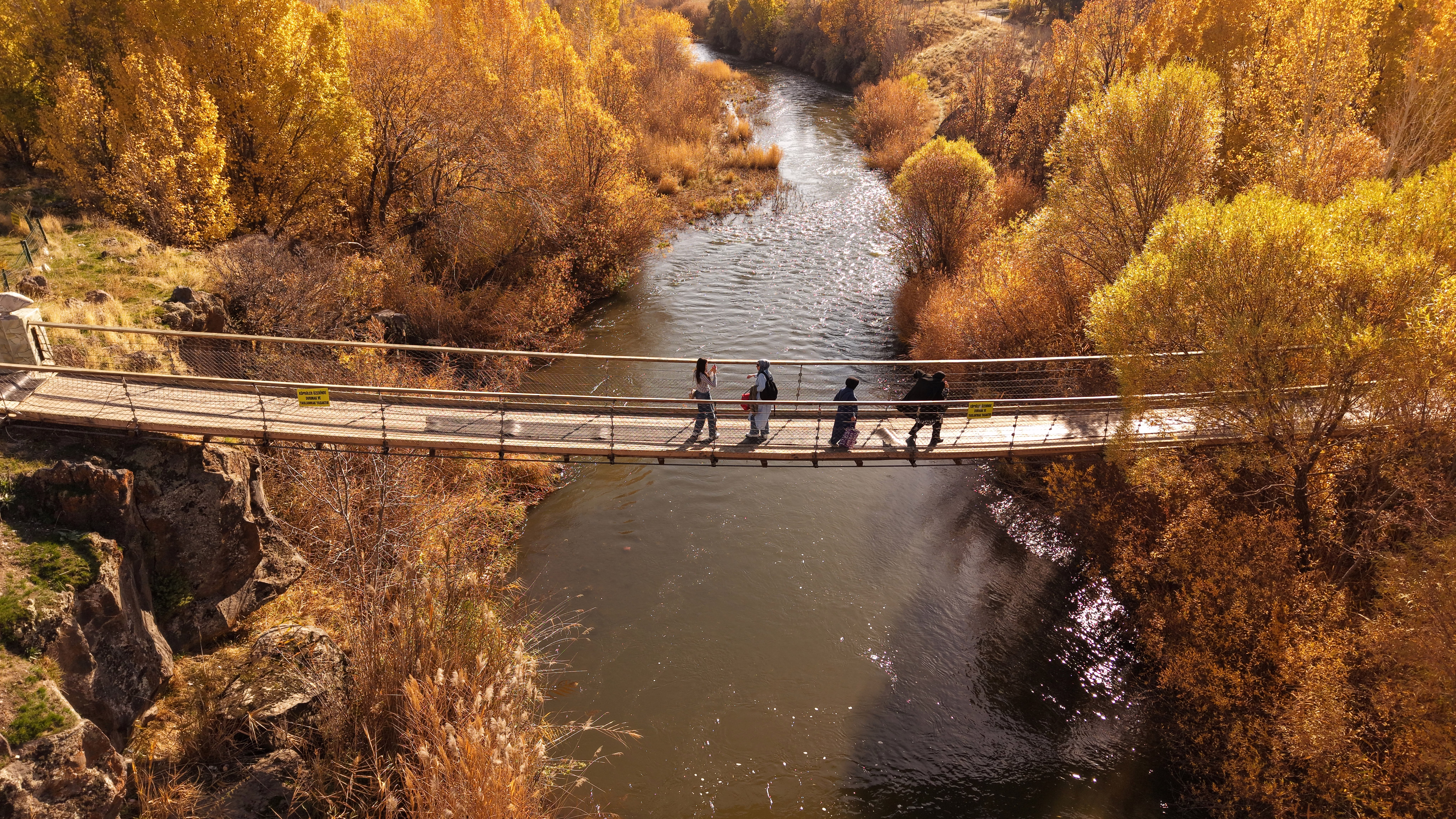 Several people walk on a foot bridge over a river, surrounded by trees in yellow fall colors.
