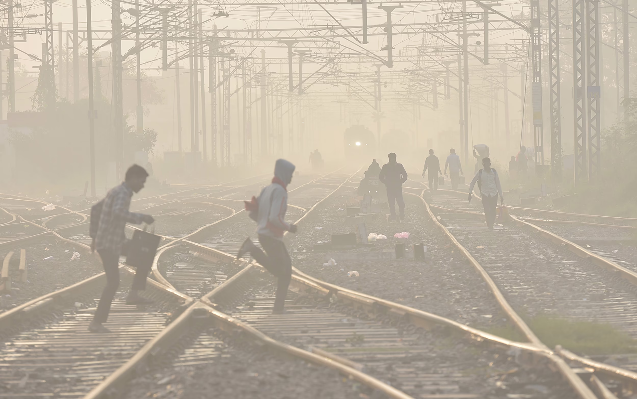 People walk through a rail yard, crossing tracks on a smoggy day.