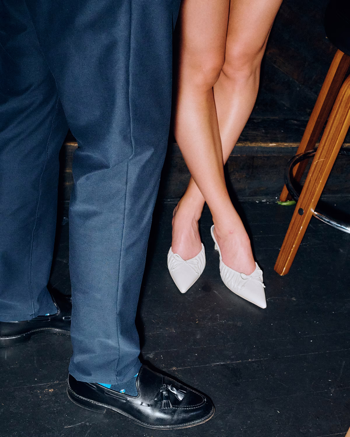 A color photograph of a woman's legs wearing white pointy heels, next to a man's legs in a blue suit.