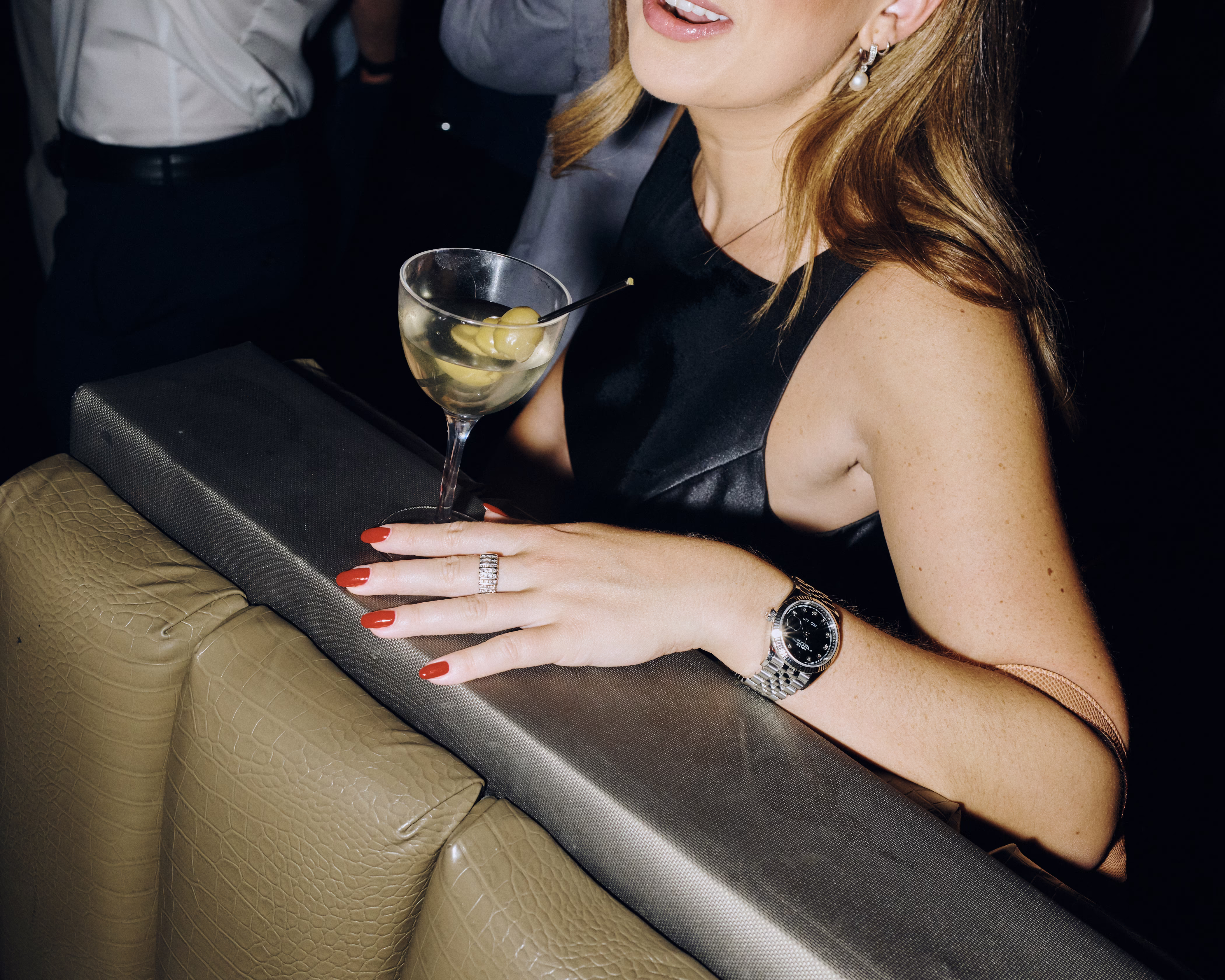 A color photograph of a woman in a black dress with red nails holding a martini