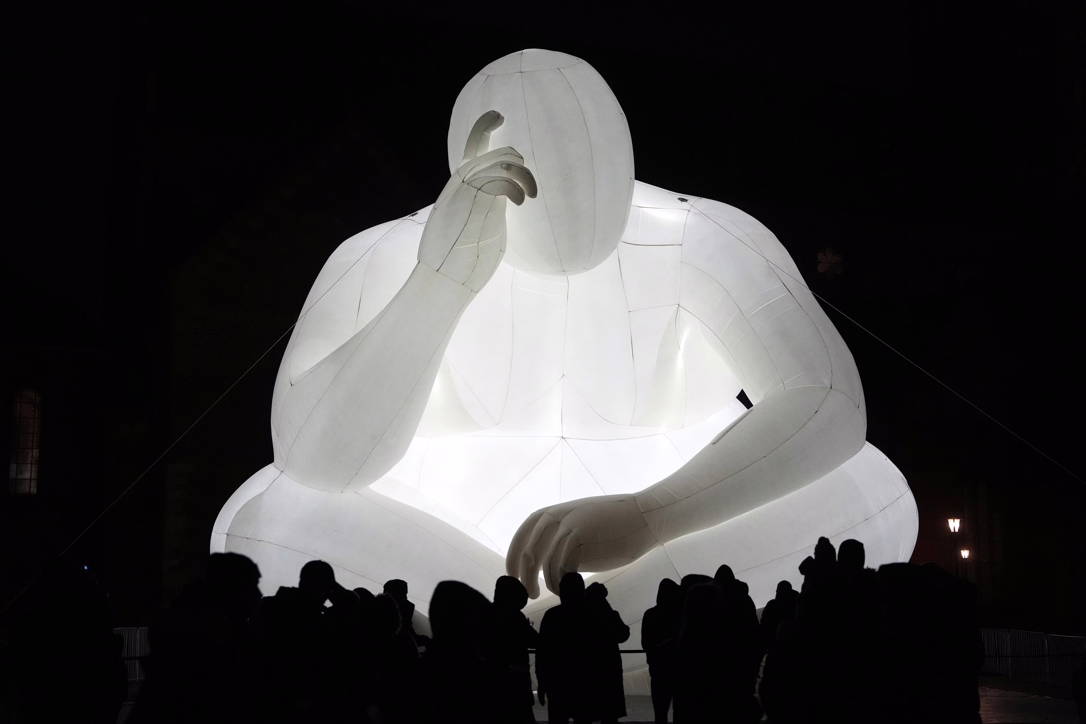 An illuminated inflatable structure shaped like a large person sitting cross-legged on the ground