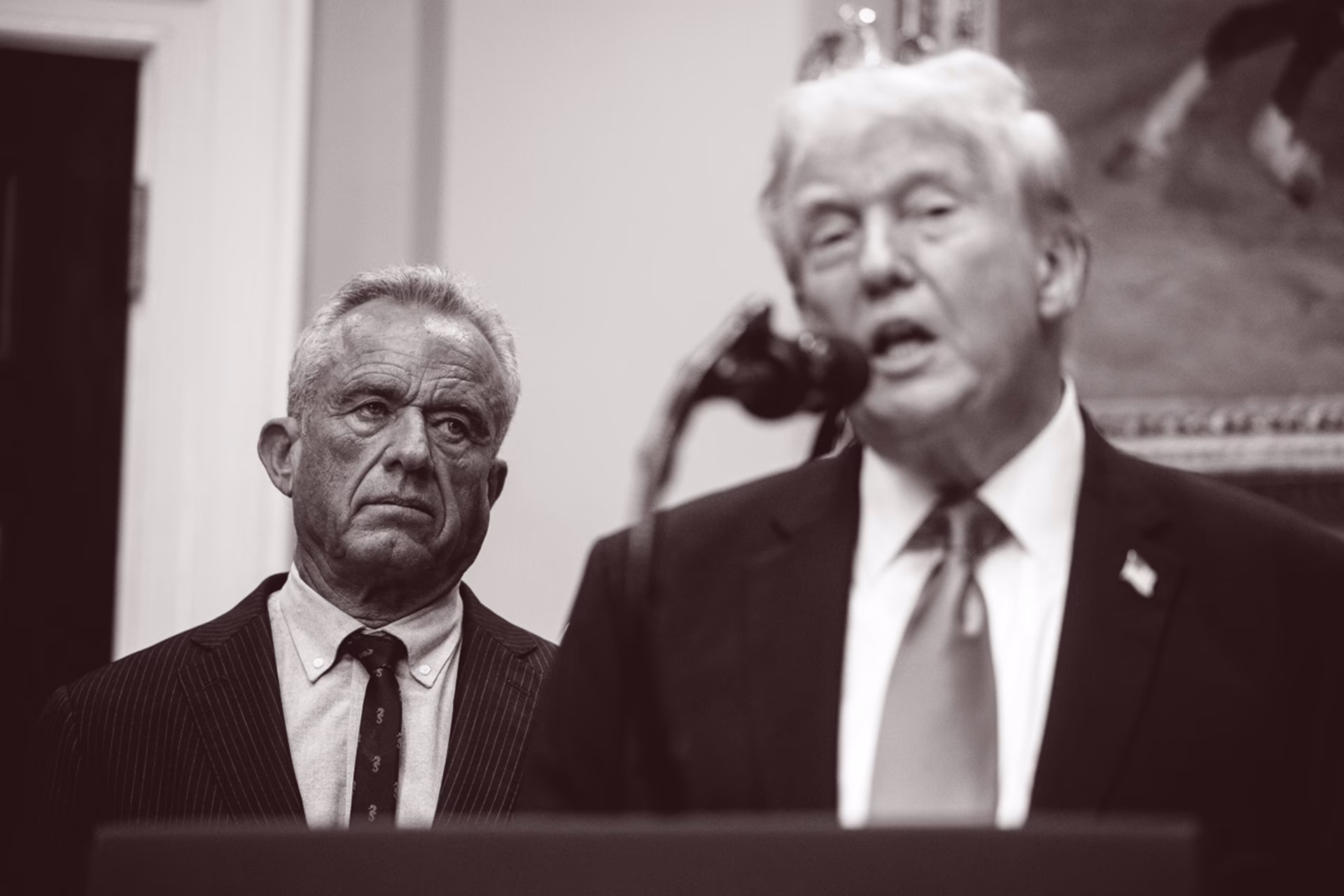 black-and-white photo of Trump speaking into microphone at podium with RFK Jr. looking on from background