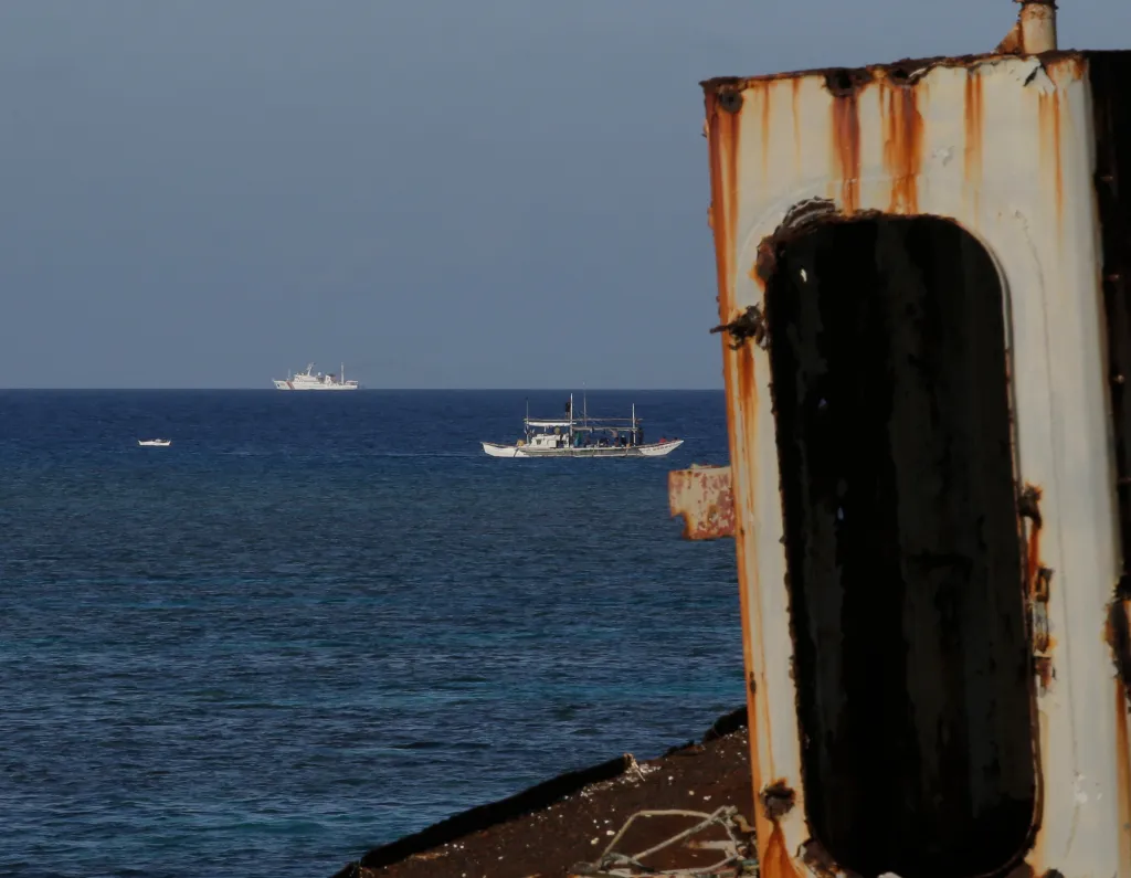 A rusted ship hull in the foreground, with three vessels at sea and a distant coastline.