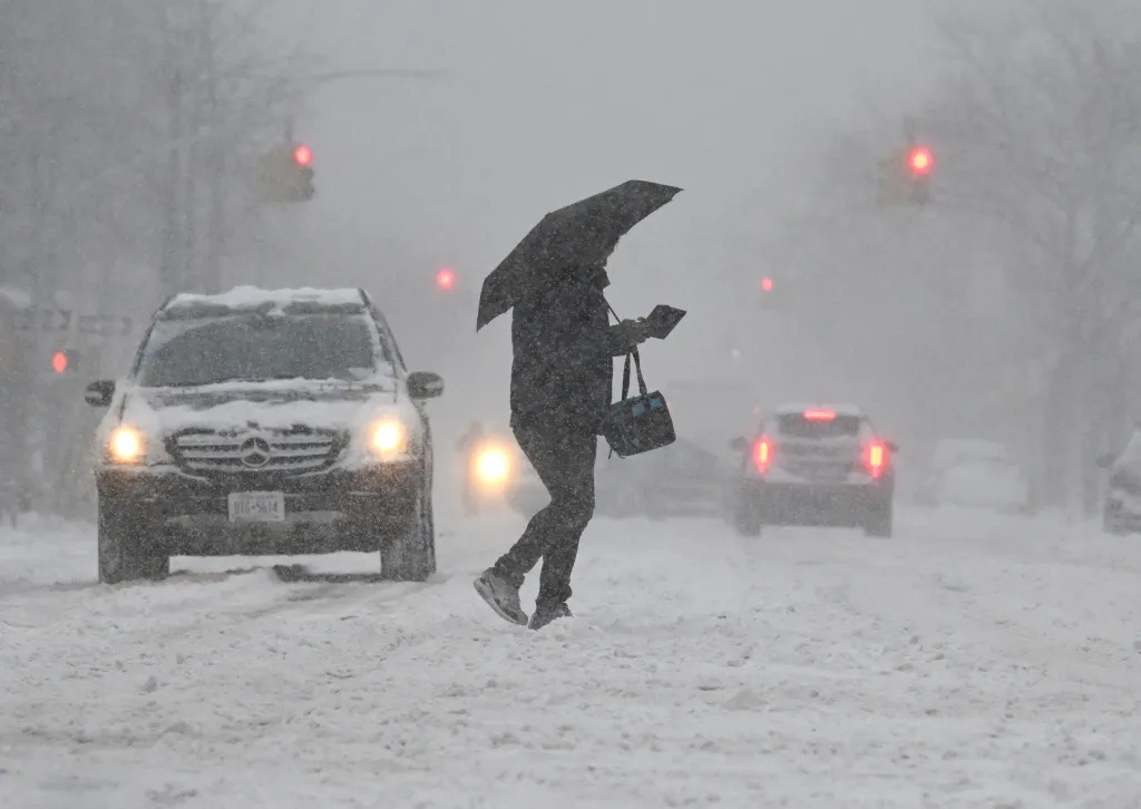 A person with an umbrella and a bag crosses a snowy street during Winter Storm Kenan.