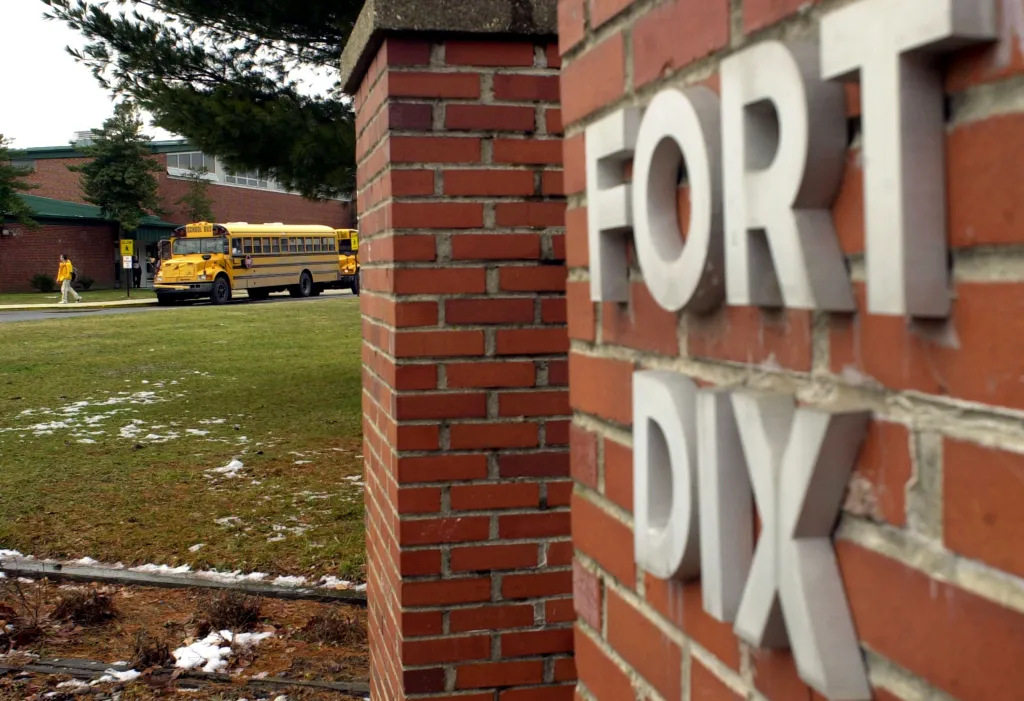 A student in a yellow jacket leaving a school building with a yellow school bus parked outside, next to a brick wall with 