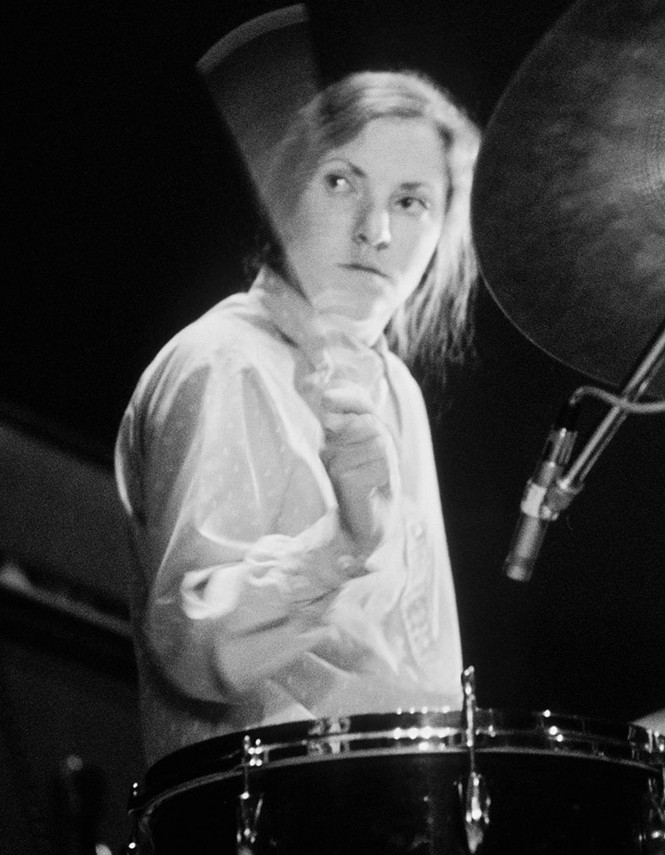 black-and-white photo of woman performing on drums with drumstick in blurred motion near her face