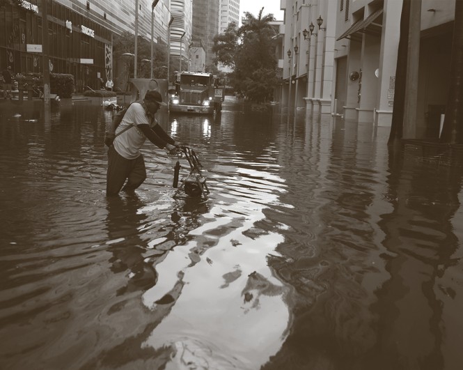 black-and-white photo of man pushing a walker through knee-deep standing water on a downtown street with tall buildings and a tow truck