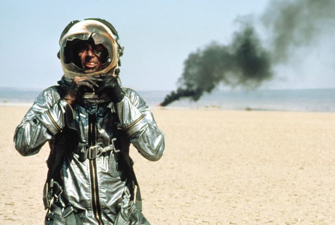 movie still of Shepard in silver test-flight suit and helmet with smashed visor walking away from smoking wreck in desert
